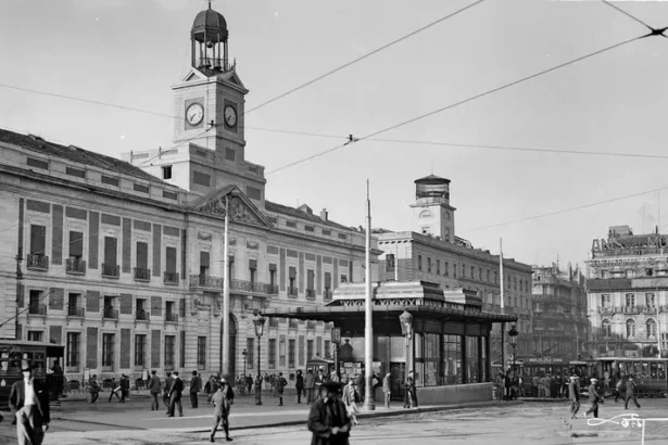 Fotografía en blanco y negro de la Puerta del Sol en 1940. El edificio es prácticamente igual que el actual, simplemente cambia la distribución de la boca del metro, más centrada frente al edificio y con una forma rectangular con tejado y ventanales para la salida.